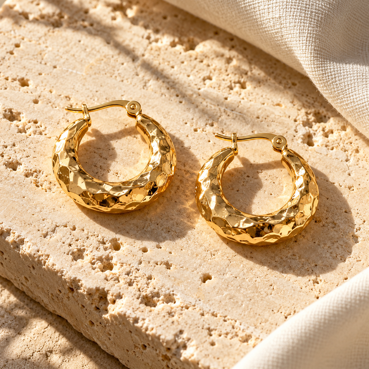 Pair of Amani textured gold hoops resting on pale sandstone